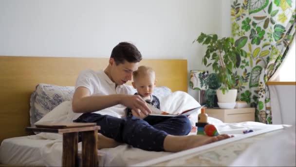 Un padre joven y su hijo pequeño leyeron un libro para niños sentado en una cama en casa — Vídeo de stock