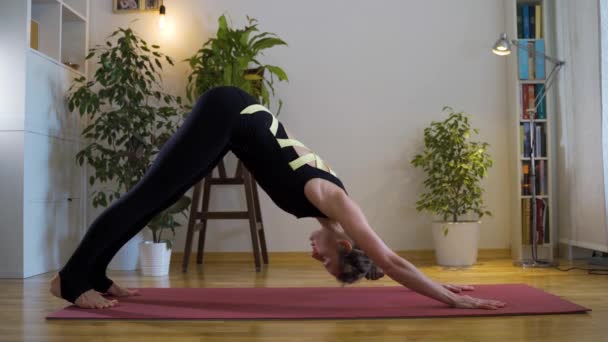 Mujer joven haciendo yoga en una alfombra roja contra una pared blanca con plantas de interior — Vídeo de stock