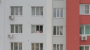 Genç bir erkek ve maskeli bir kadın ailesi pencereden dışarı bakar, salgın sırasında karantinada kalırlar. Hava, kamera dışarı.