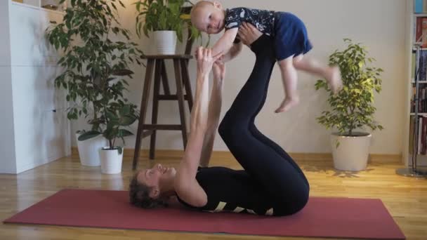 Jeune femme faisant du yoga avec son enfant sur un tapis rouge contre un mur blanc avec des plantes d'intérieur — Video