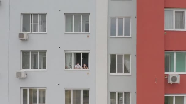 En ung familj till en man och en maskerad kvinna tittar ut genom fönstret, stannar hemma i karantän under pandemin. Flyg, kameran rör på sig. — Stockvideo