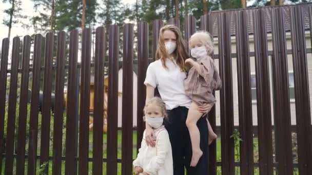 A young mother and two young daughters in mask stand at a fence outside a house in the village during a quarantine pandemic — Stock Video