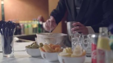 Siyah ceketli ve beyaz gömlekli barmen sisli bir fıçıdaki buzu metal bir çalkalayıcıya koyar ve kapatır. Masanın üzerinde yeşil limon dilimleri, turuncu, sarı limon ve bardaklar var.