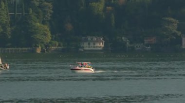 Neşeli renkli renkli balonlarla süslenmiş bir motorlu tekne, güneşli bir yaz günü Como Gölü 'nde beyaz bir yatın arkasında yeşil ağaçların ve İtalyan evlerinin önünde yüzüyor. Orta plan