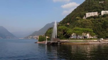 Güneşli bir yaz sabahında Villa Geno 'daki çeşmenin güzel manzarası. Como Gölü 'nün kıyısındaki bir drondan ateş ediliyor. İnsanlar rahatlar, patika boyunca yürürler, egzotik ağaçların yanında güneşin altında otururlar.