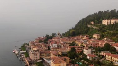İtalya 'da Como Gölü' nde Lombardy 'de güzel bir şehir olan Bellagio' nun hava manzarası. Yeşil ağaçların arasında sarı, beyaz, kırmızı İtalyan evleri. Bulutlu bir yaz gününde bir gemi rıhtımda duruyor.
