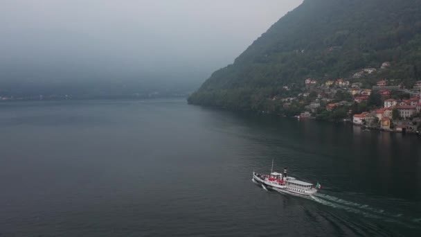 白い小さなヴィンテージのクルーズ船は 曇った夏の日に山の中腹に緑の木の真ん中にイタリアの家を背景にコモの青い湖を航海します ドローンからの空中の眺め — ストック動画