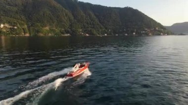 Kırmızı motorlu teknedeki yeni evliler, Como 'nun mavi gölü boyunca hızla ilerleyip arkalarında büyük dalgalar bırakarak yaz günbatımında yeşil dağların arka planına doğru ilerliyorlar. Hava görünümü