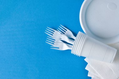 Tek kullanımlık plastik sofra takımı, çatallar, bardaklar, mavi arka planda plakalar. Sorun geri dönüşüm, yeniden kullanım, güvenli gezegen, çevre kavramı. Metin için yer.