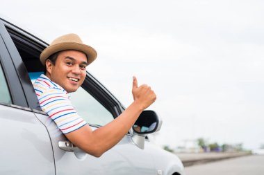 Genç Asyalı adam araba sürerken baş parmağını gösteriyor..