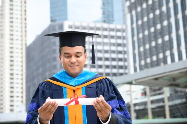 Happy graduate. Happy Asain man in graduation gowns holding diploma in hand on urban city background.