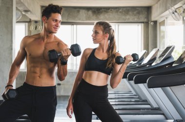 Fitness adamı ve kadın elinde dambıl tutarak koşu bandının yanında poz veriyorlar. Spor salonunda kaslı çift eğitimi..