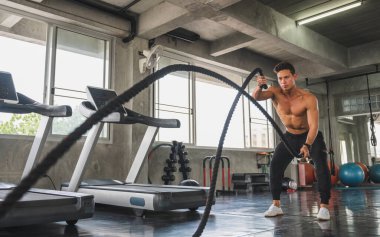 Genç adam spor salonunda savaş halatlarıyla çalışıyor..