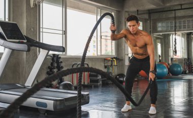 Genç adam spor salonunda savaş halatlarıyla çalışıyor..