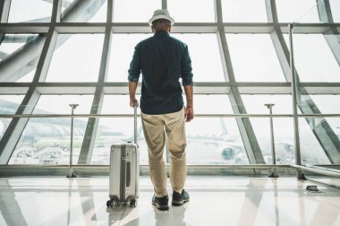 Seyahate çıkmaya hazırlanan gri şapkalı bir erkek yolcu. Bir tramvay çantası var. Dünyayı dolaşmak için büyük bir havaalanında duruyor..