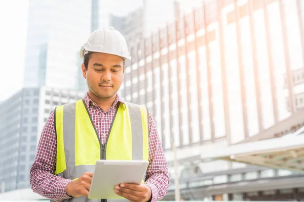 Young handsome asian civil engineer holding tablet looking forward and thinking, planning on construction building background. Evolution construction concept.