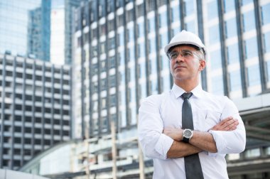 İnşaat alanının arka planında düşünme, gülümseme ve kol çapraz sorgulaması yapan kıdemli inşaat mühendisinin portresi.