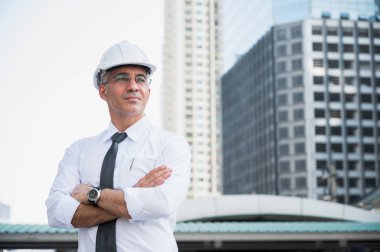 İnşaat alanının arka planında düşünme, gülümseme ve kol çapraz sorgulaması yapan kıdemli inşaat mühendisinin portresi.