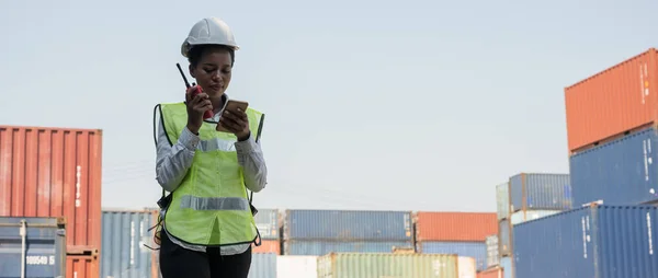 Black foreman woman worker working checking at Container cargo harbor holding radio walkie-talkie and smartphone to loading containers. African dock female staff business Logistics import export shipping concept.