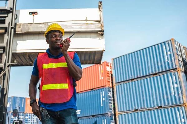 Black foreman worker working control the crane and forklift at Container cargo harbor to loading containers. African dock male staff with radio walkie-talkie for Logistics import export shipping concept.