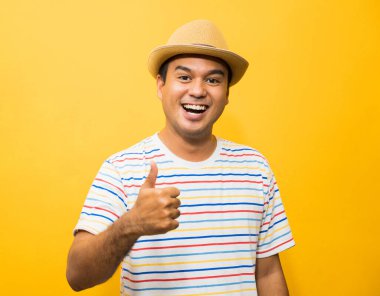 Şapkalı genç Asyalı adam mutlu ve şaşırmış hissediyor ve sarı arka planda başparmağını gösteriyor..