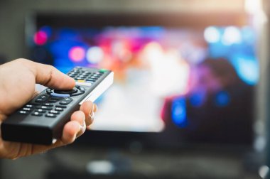 Elinde televizyon kumandası olan genç bir adam. Eller televizyon ekranını işaret ediyor ve onu açıyor ya da kapatıyor. Oturma odasındaki kanepede televizyon seyrediyor. Rahatla..
