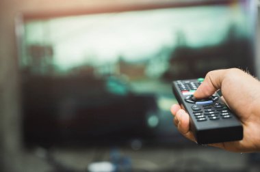 Elinde televizyon kumandası olan genç bir adam. Eller televizyon ekranını işaret ediyor ve onu açıyor ya da kapatıyor. Oturma odasındaki kanepede televizyon seyrediyor. Rahatla..