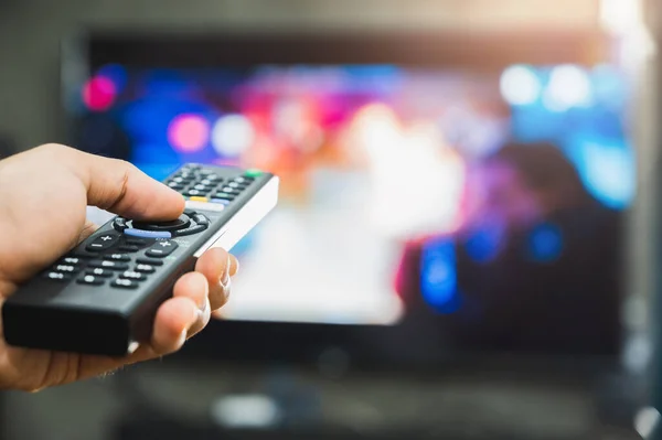 Elinde televizyon kumandası olan genç bir adam. Eller televizyon ekranını işaret ediyor ve onu açıyor ya da kapatıyor. Oturma odasındaki kanepede televizyon seyrediyor. Rahatla..