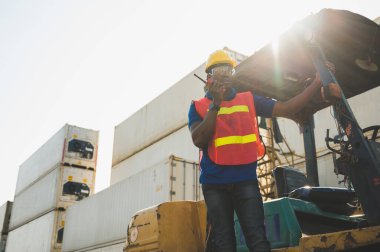Kara ustabaşı konteynır kargo limanındaki vinç ve forklifti kontrol ediyor. Lojistik ihracat nakliye konsepti için telsiz telsiziyle Afrika rıhtımı erkek personeli.