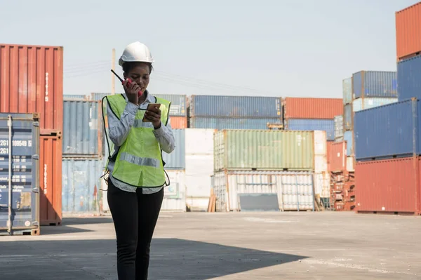 Black foreman woman worker working checking at Container cargo harbor holding radio walkie-talkie and smartphone to loading containers. African dock female staff business Logistics import export shipping concept.