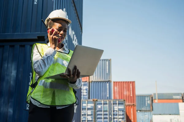 Black foreman woman worker working checking at Container cargo harbor holding laptop computer and walkie-talkie to loading containers. African dock female staff business Logistics import export shipping concept.