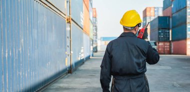 Ustabaşı işçi, konteyner kargo limanında çalışıyor. Elinde telsiz telsiziyle konteynırları yüklüyor. Rıhtım erkek personel lojistik ihracat nakliyat konsepti.