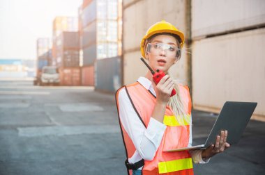 Ustabaşı kadın işçi, konteyner kargo limanında dizüstü bilgisayarı ve telsiz telsiziyle konteynırları kontrol ediyor. Rıhtım kadın personel lojistik ithalat ihracat nakliye kavramı.