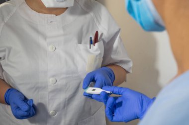 Hemşire termometreyi başka bir hemşireye veriyor. Hemşireler mavi eldiven ve yüz maskesi takarlar. Yüksek ateş koronavirüsün semptomlarıdır..