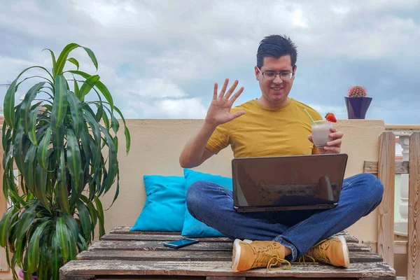 Man waving for a virtual call with his computer. He is on the terrace of his house. It has a strawberry juice.