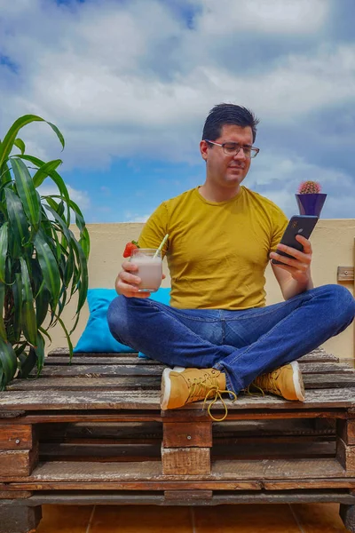  Evinin balkonunda cep telefonuna bakan bir adam. Çilekli smoothie içiyor..