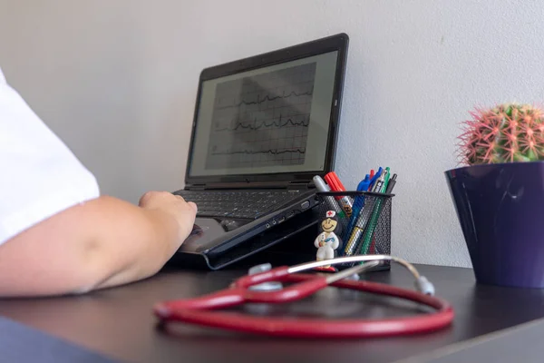 On a desk in the foreground a stethoscope. In the background is a pencil with pens, a cactus and a laptop. All out of focus.
