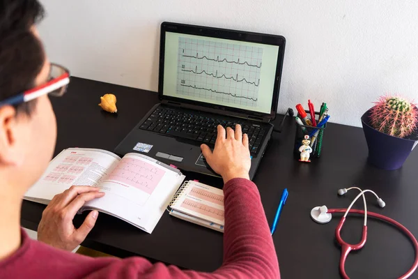 Young man studying English sitting at a desk. On the desk is a laptop with an electrocardiogram on the screen. There\'s also a cardiology textbook. On the desk there is a stethoscope and a cactus.