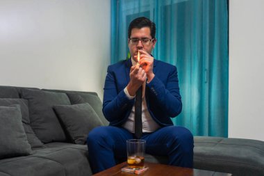 Kanepede oturan genç adam. O mavi bir takım elbise giyiyor. O bir puro yakıyor. Tahta bir masanın üzerinde bir bardak viski var..