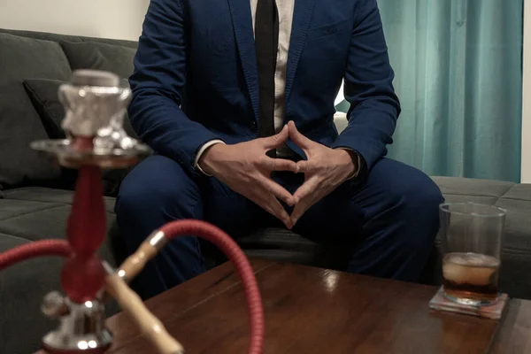 Young man in a blue suit sitting on a sofa. He is smoking a hookah and drinking a glass of Whiskey.