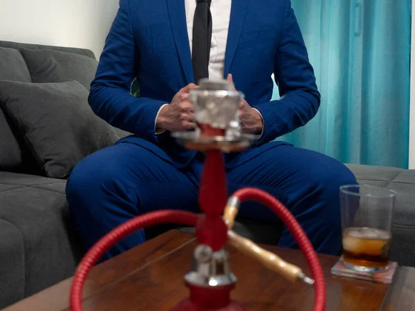 Young man in a blue suit sitting on a sofa. He is smoking a hookah and drinking a glass of Whiskey.