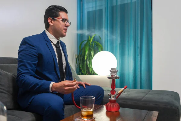 Kanepede oturan genç adam. O mavi bir takım elbise giyiyor. O bir bardak viski içiyor. Tahta bir masanın üzerinde kırmızı bir nargile var..