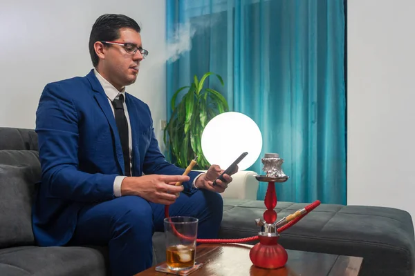 Kanepede oturan genç adam. O mavi bir takım elbise giyiyor. O, cep telefonuna bakıyor. Tahta bir masanın üzerinde bir bardak viski ve kırmızı bir shisha var..