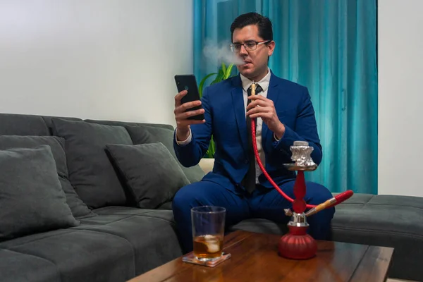 Kanepede oturan genç adam. O mavi bir takım elbise giyiyor. O, cep telefonuna bakıyor. Tahta bir masanın üzerinde bir bardak viski ve kırmızı bir shisha var..