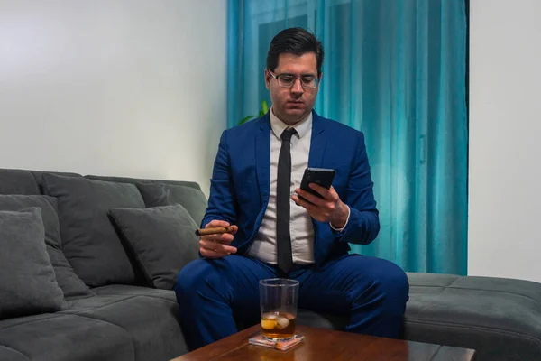 Young man in a blue suit sitting on a sofa. He is smoking a cigar and drinking a glass of Whiskey. He is looking at his mobile phone.