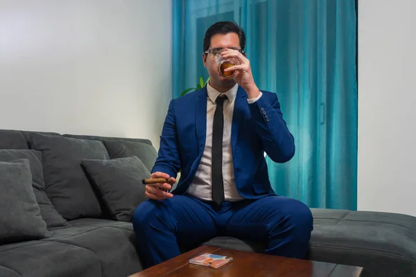 Young man in a blue suit sitting on a sofa. He is smoking a cigar and drinking a glass of Whiskey.