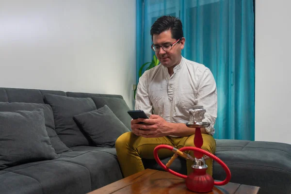 Kanepede oturan genç adam. Cep telefonuna bakıyor ve bir shisha ile sigara içiyor..