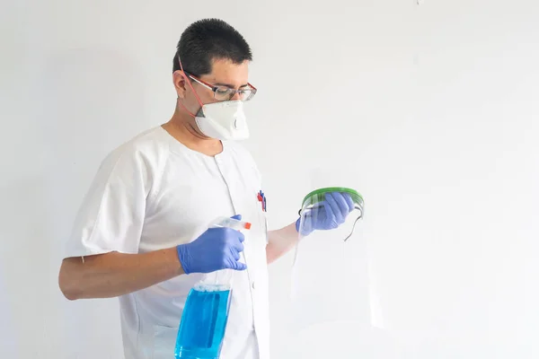 Doktor / hemşire yüz kalkanını temizliyor. Eldiven ve maske takıyor. Üniforması beyaz..
