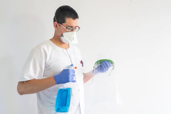 Doktor / hemşire yüz kalkanını temizliyor. Eldiven ve maske takıyor. Üniforması beyaz..