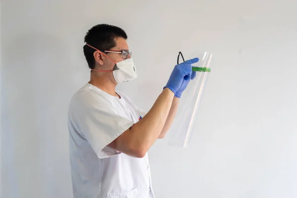 Doktor / hemşire, koronavirüsten korunmak için yüz kalkanı takıyor. O genç bir adam. Eldiven, maske ve gözlük takıyor. Beyaz bir üniforma giyiyor..